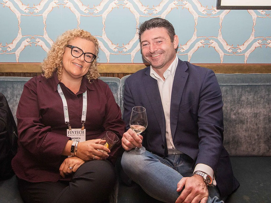 Two guests seated on a velvet banquette with drinks, smiling at the camera