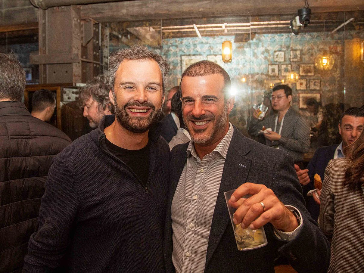 Two guests smiling together in a busy, lamp-lit bar
