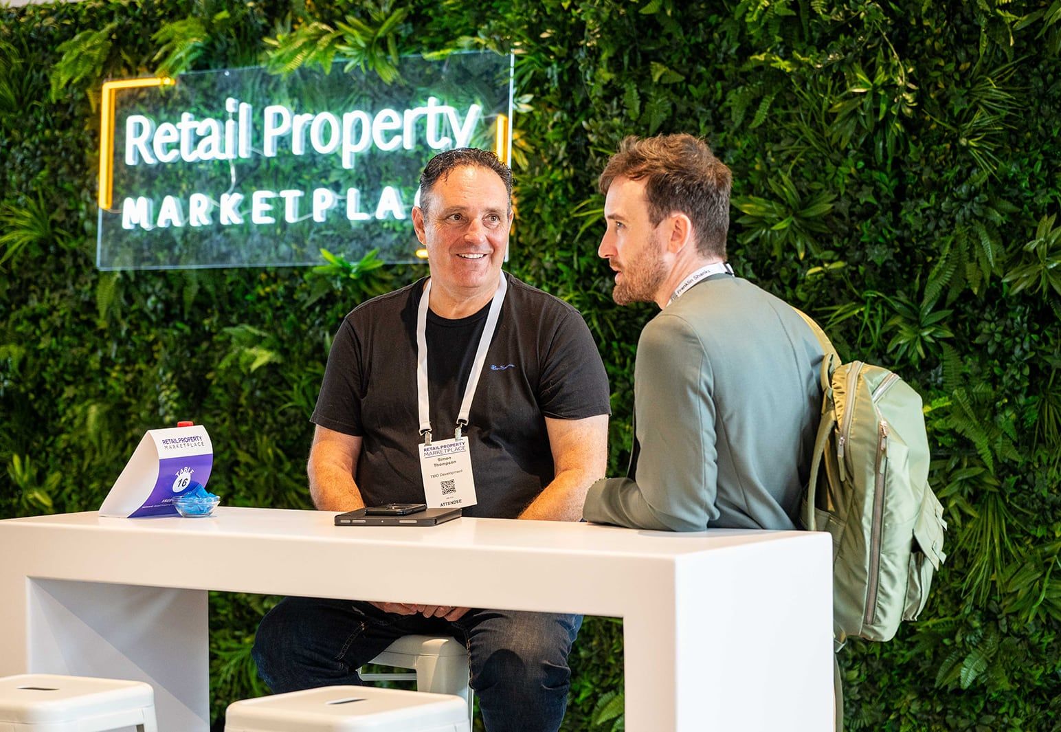 Two people meeting at a table in front of the Retail Property Marketplace sign on a green wall