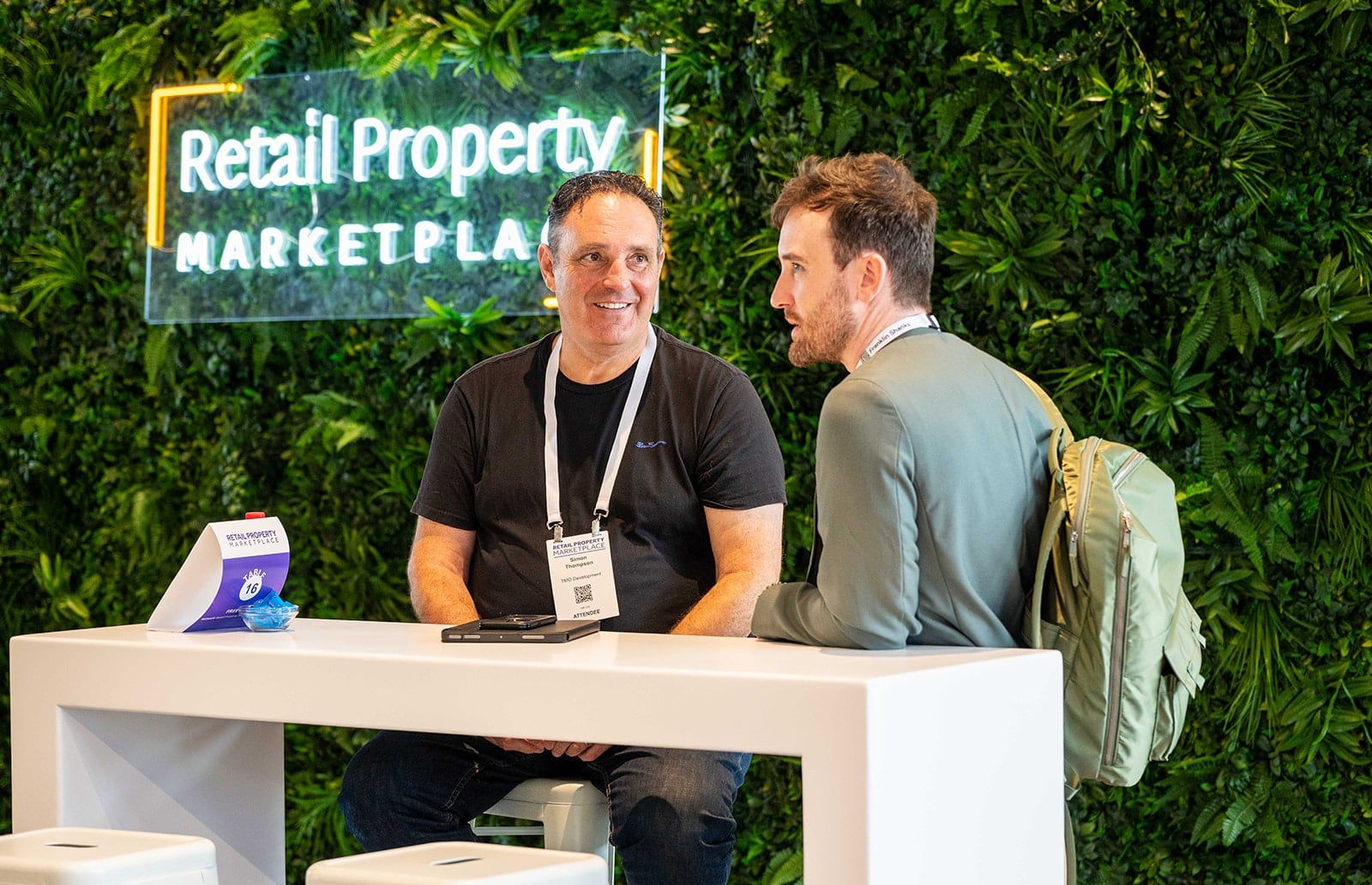 An exhibitor in a dark jacket explaining something to two visitors, with plants behind them