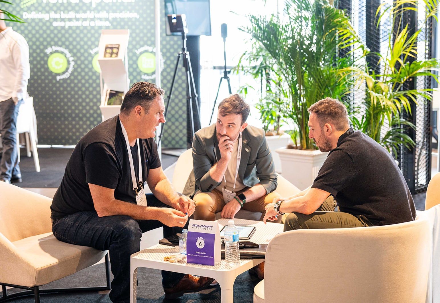 Three people in conversation around a low table on lounge chairs, with palms behind them