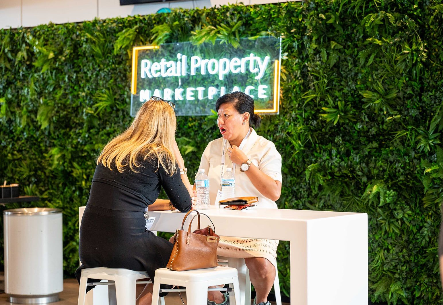 Two women meeting at a table below the Retail Property Marketplace neon sign
