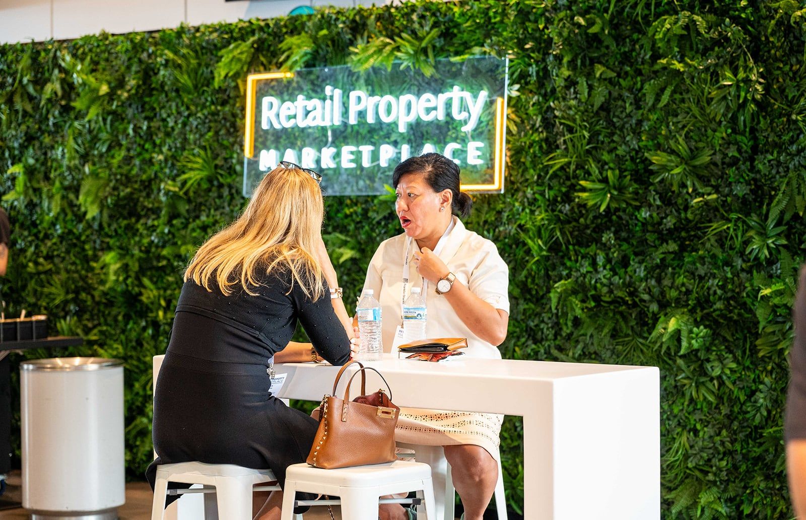 Two women meeting at a table below the Retail Property Marketplace neon sign