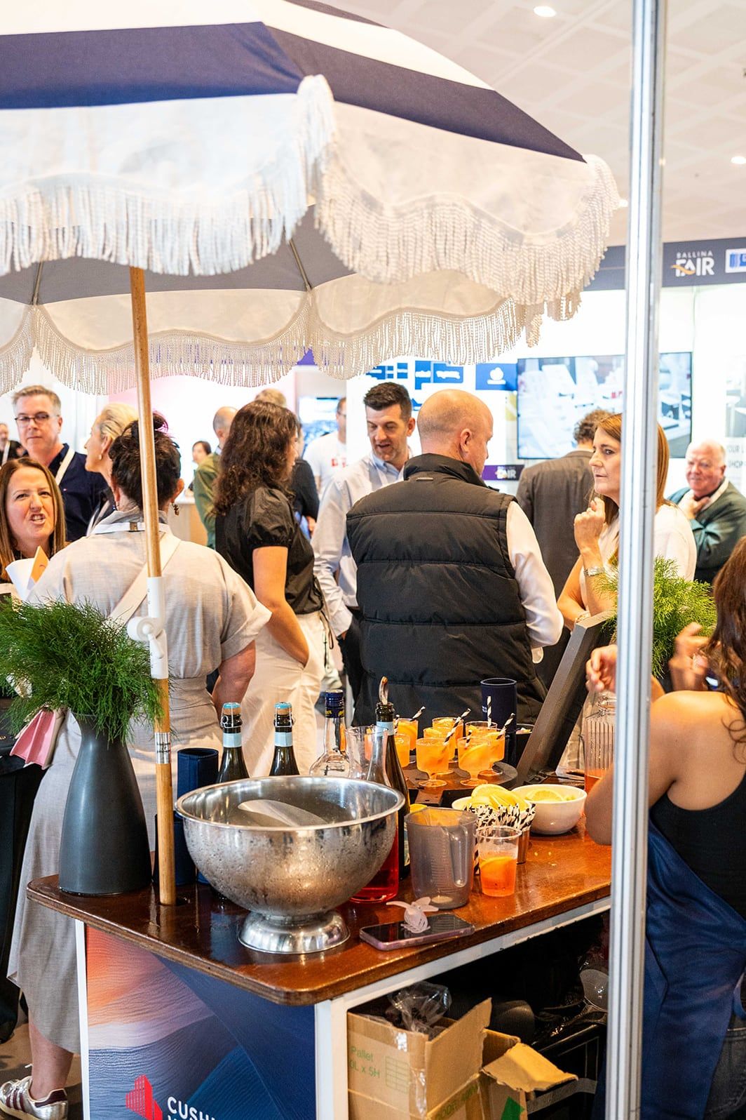 Guests gathered at a drinks station under a striped umbrella on the exhibition floor