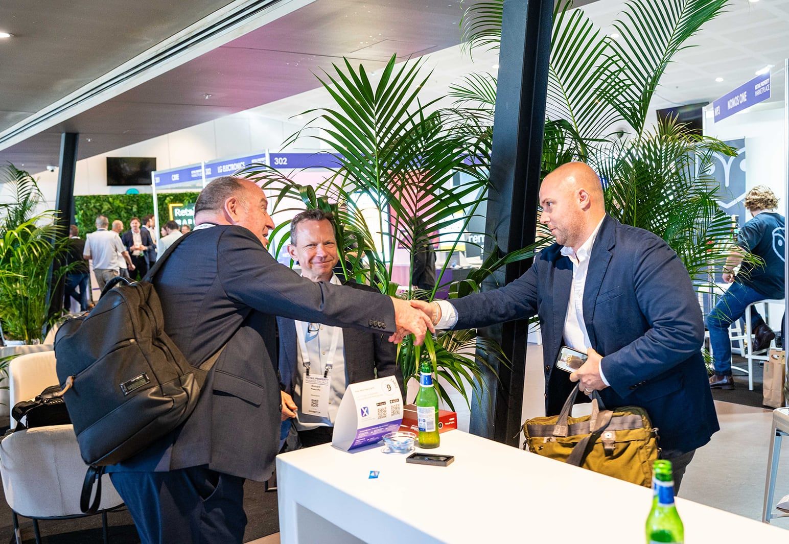 Two men shaking hands at a white table among palms while a third looks on