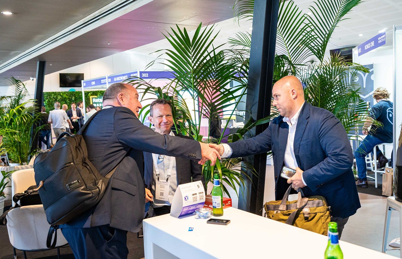 Two men shaking hands at a white table among palms while a third looks on