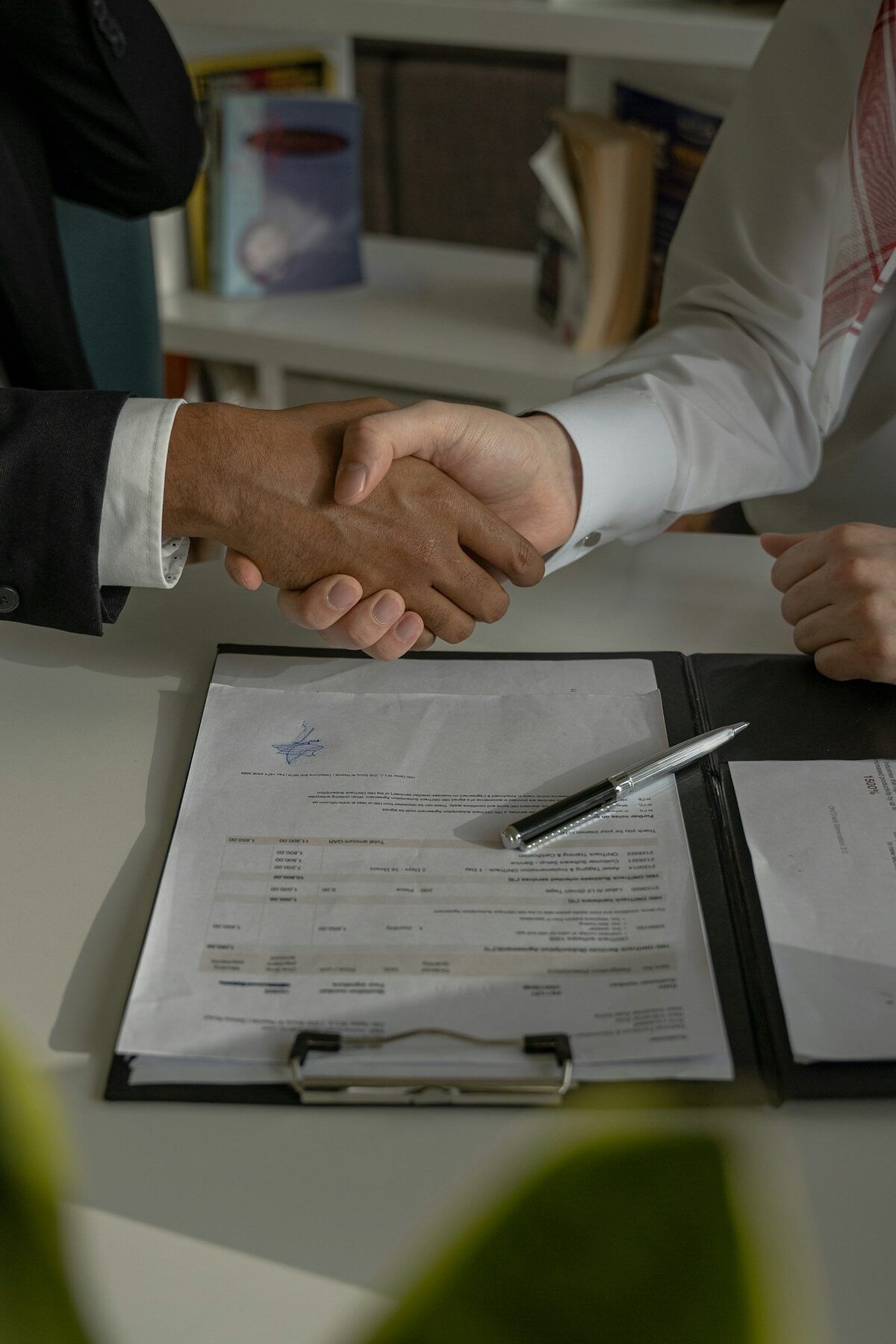 Two people shaking hands over a signed contract and pen on a clipboard