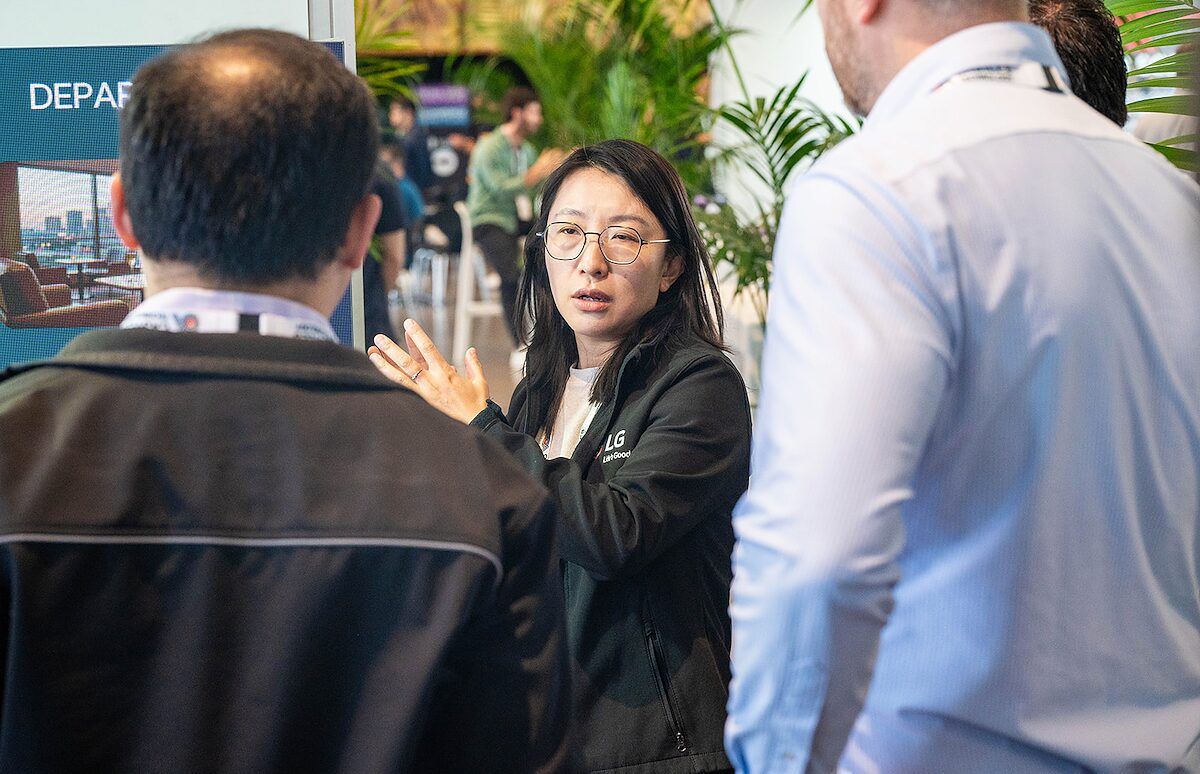 An exhibitor in a dark jacket explaining something to two visitors, with plants behind them
