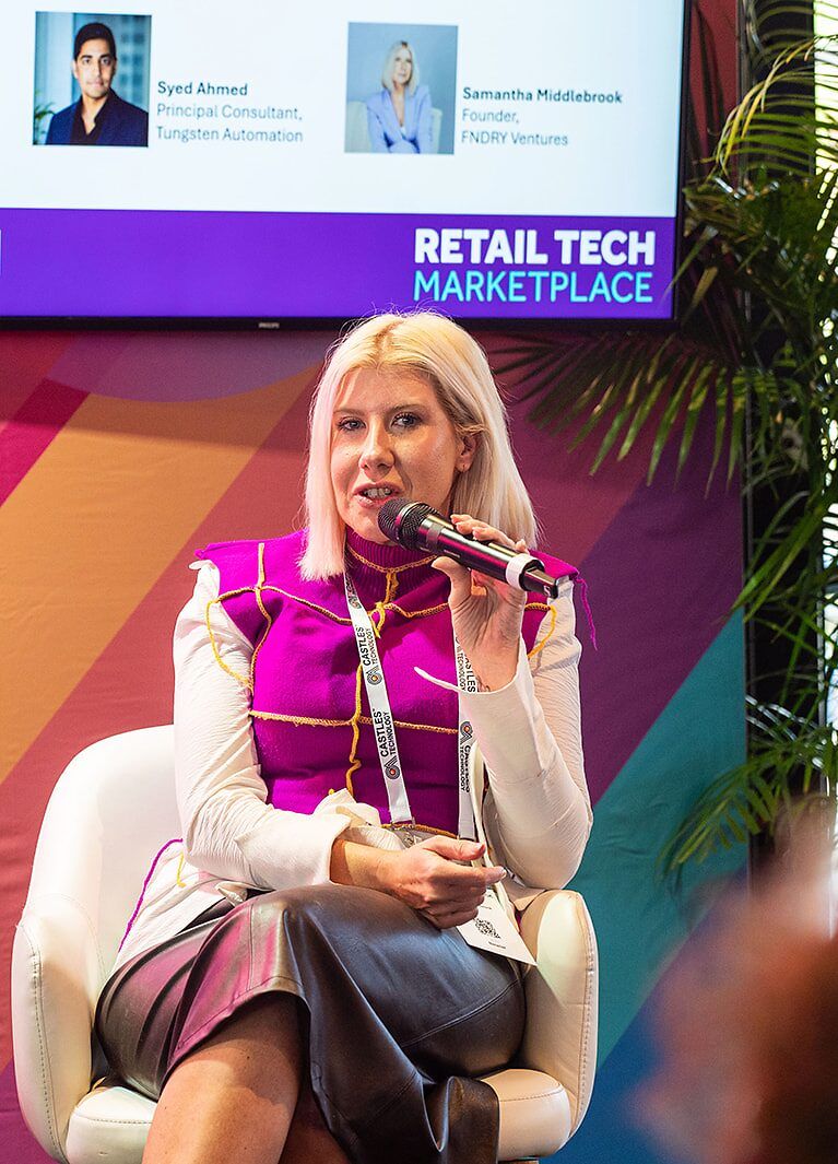 A speaker with a microphone seated on the Retail Tech Marketplace stage, with the session screen behind her