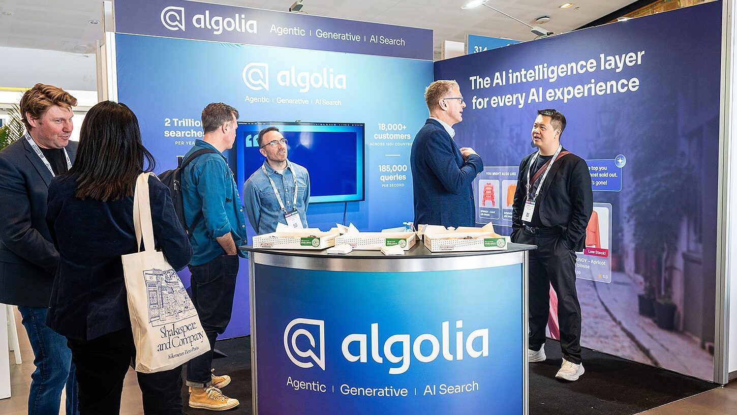 Two exhibitors and a visitor talking at a blue branded stand with a round counter and a large screen