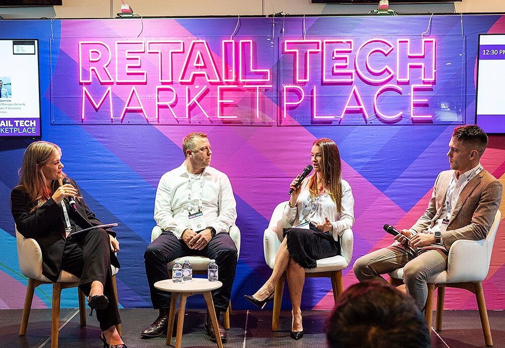 A panel of four seated on the Retail Tech Marketplace stage, with session screens on either side