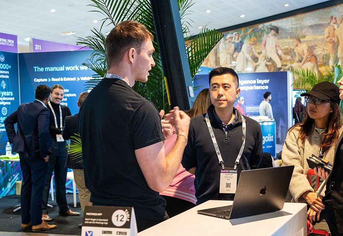 An exhibitor talking with visitors across a stand counter with a laptop, among palms