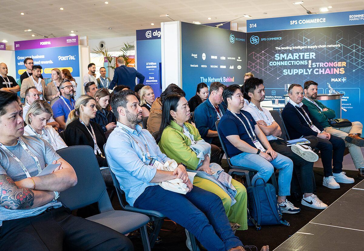 A seated audience watching a session on the event floor, with stands behind them