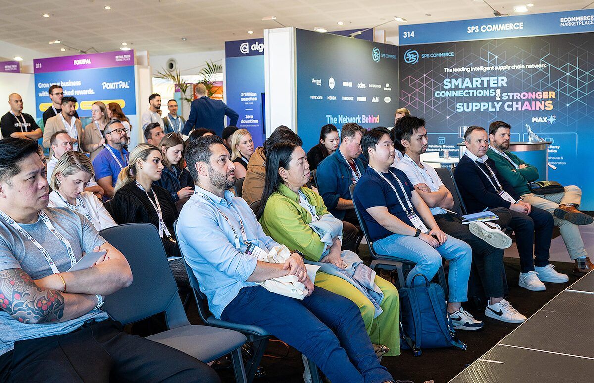 An audience seated in front of a stage, with exhibitor stands in the background