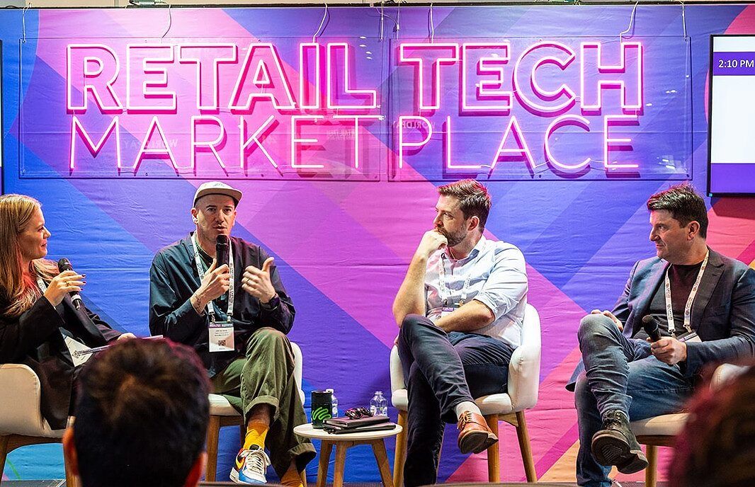 Three speakers in conversation with a host on the Retail Tech Marketplace stage, lit by a neon sign