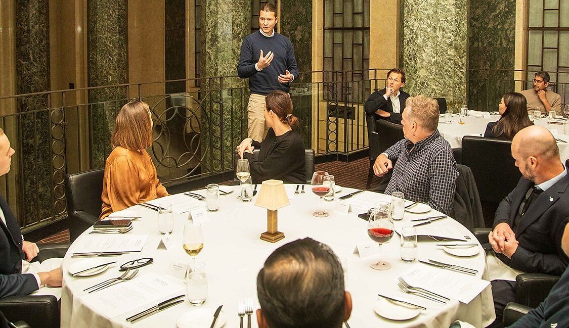 Guests seated around a set dinner table listening to a speaker at an executive dinner