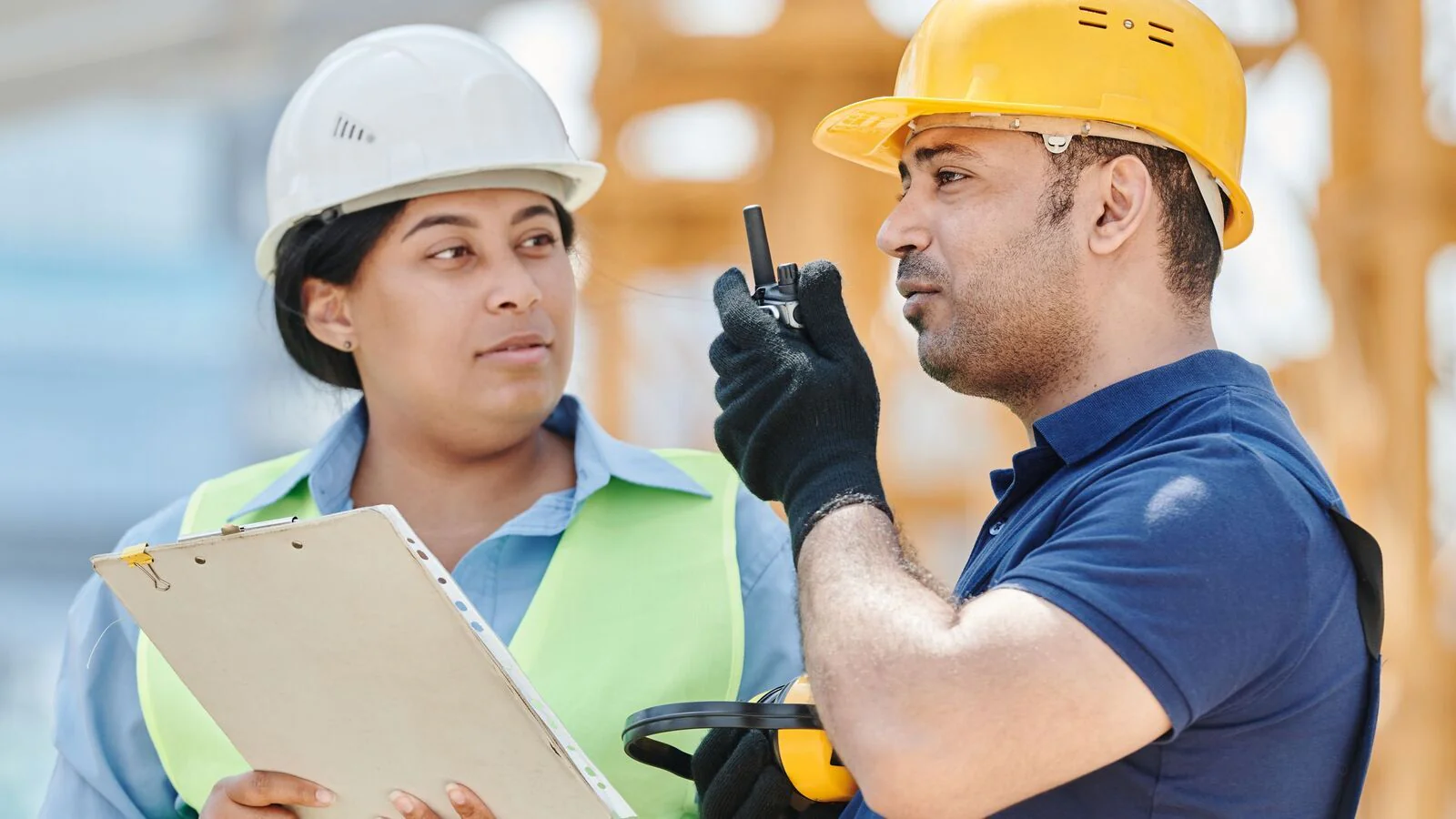Two site workers in hard hats, one with a clipboard and one on a two-way radio
